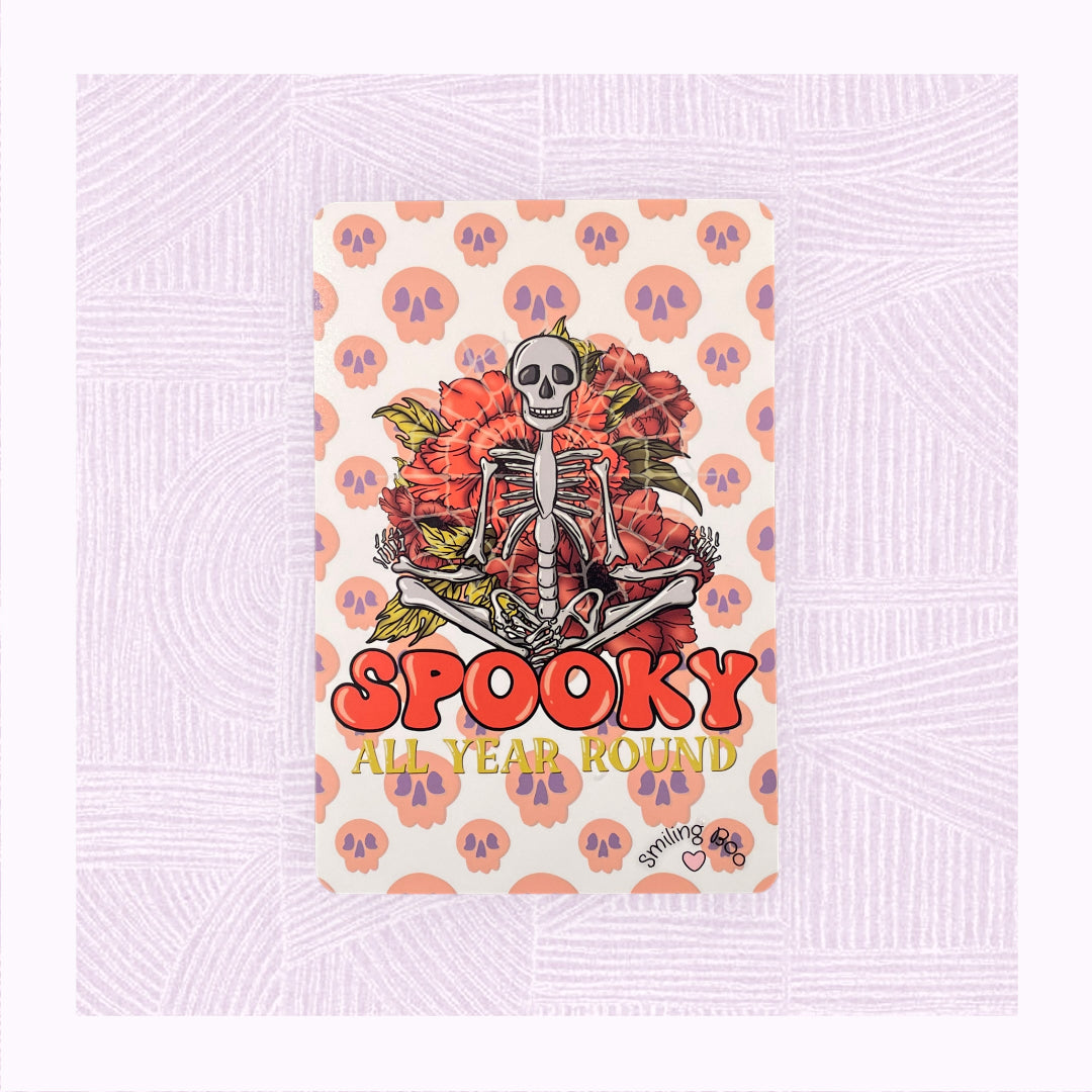 Postcard art print with a background of raining skulls, a large skeleton in the foreground with the phrase ‘Spooky All Year Round’ underneath it.