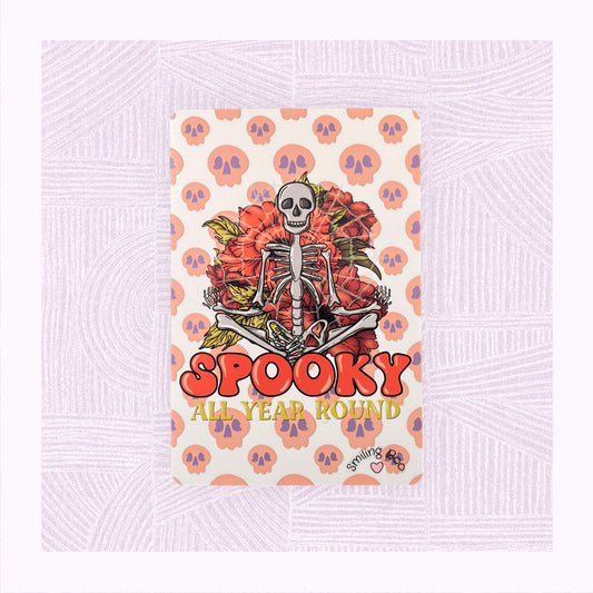 Postcard art print with a background of raining skulls, a large skeleton in the foreground with the phrase ‘Spooky All Year Round’ underneath it.
